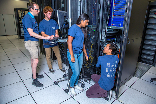 A mentor and three students work alongside the open doors of an HPC cluster's cabinet
