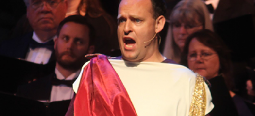Jarom Nelson performing in Roman dress in front of a choir of formally dressed singers