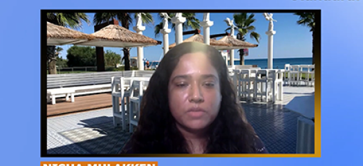 Screen shot of Nisha speaking to the camera with a virtual background behind her