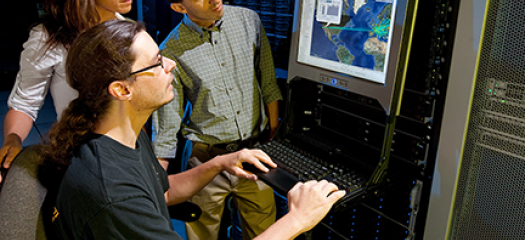 three people look at a computer screen attached to a supercomputer