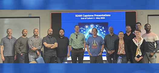 the first cohort of SEAM participants stand as a group in front of a large video screen