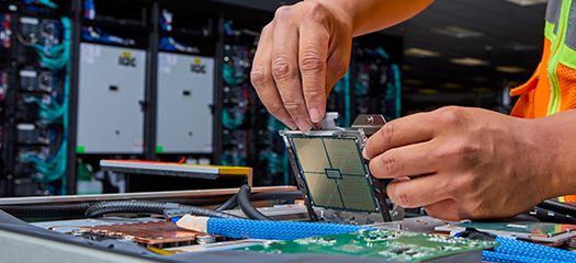 a person’s hands are shown installing a computer chip into a flat section of a supercomputer with a cabinet from the full system in the background