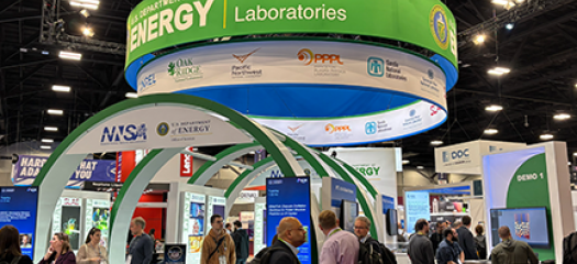 people clustered in and around the Department of Energy booth, which consists of large green and white circular structures with a demo station on the right side