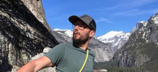 Man in baseball cap and outdoor gear leaning against the side of a large rock outdoors in a mountain range