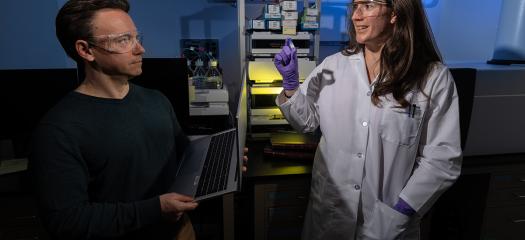 Colin holding a laptop and Carolyn holding a sample; they are standing in a lab