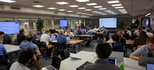 dozens of people with laptops at tables in a large classroom setting