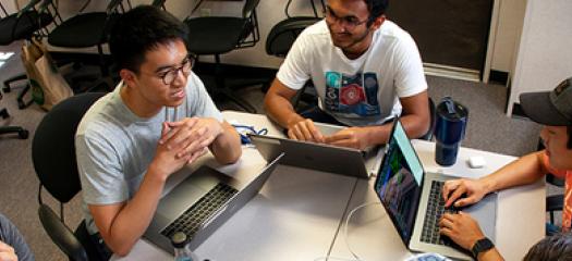 developers sit in a circle around a table, working on their laptops