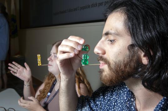 Developer Day attendees are balancing a stack of dice on popsicle sticks held in their mouths. 