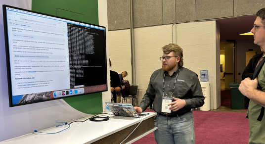 large screen on the left displaying code and GitHub instructions, Hobbs in the middle with his laptop, and viewers on the right