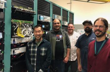 Five people are standing next to machine racks.