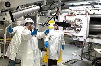 Three people in lab coats and personal protective equipment are moving large machinery.