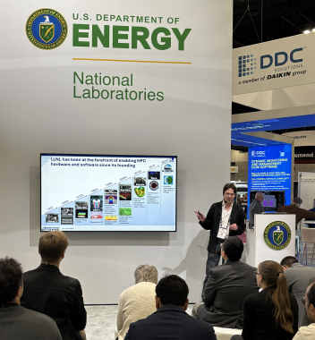 Nikoli near a podium gesturing to a timeline of the Lab’s HPC systems on screen, which is on a wall displaying the DOE logo