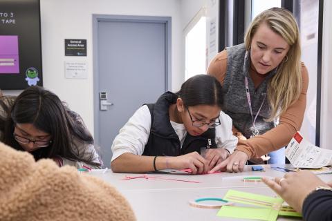 Adult leans over a student who is folding a piece of paper 