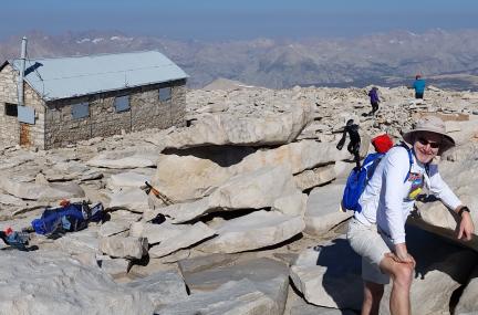 Mark Miller is leaning against a large rock and smiling on the summit plateau of Mount Whitney, wearing a white shirt, sunglasses, a blue backpack, and a hat. He is on the bottom right of the photograph, surrounded by large rocks. There is a small shelter behind him and mountains on the horizon.