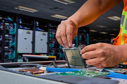 a person’s hands are shown installing a computer chip into a flat section of a supercomputer with a cabinet from the full system in the background