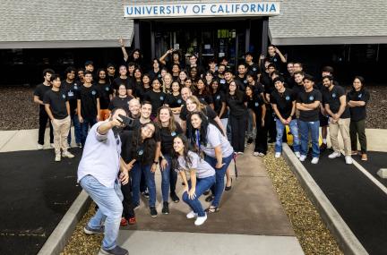 Large group is gathered  outsie of a  building with the person in front attempting to take a selfie