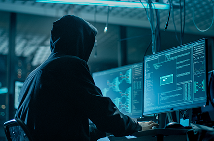 Hooded person hacking into a computer in a dark atmosphere with multiple monitors and cable