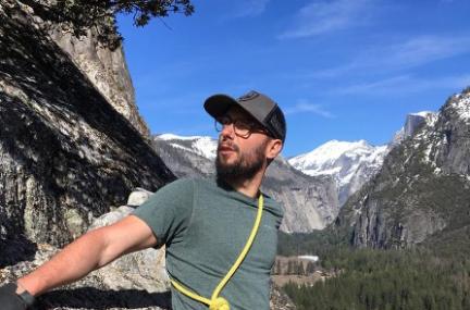 Man in baseball cap and outdoor gear leaning against the side of a large rock outdoors in a mountain range
