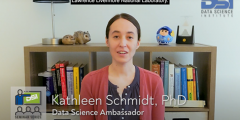 Katie stands in front of a bookcase and speaks into the camera