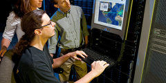 three people look at a computer screen attached to a supercomputer