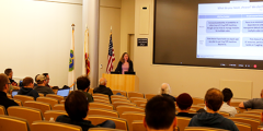 Judy Hill presents at a lectern to an auditorium of people