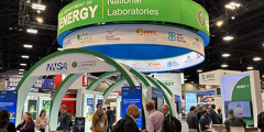 people clustered in and around the Department of Energy booth, which consists of large green and white circular structures with a demo station on the right side