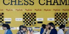  several chess players seated at tables during a competition, in front of competition-related signs on a yellow wall