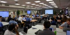 dozens of people with laptops at tables in a large classroom setting