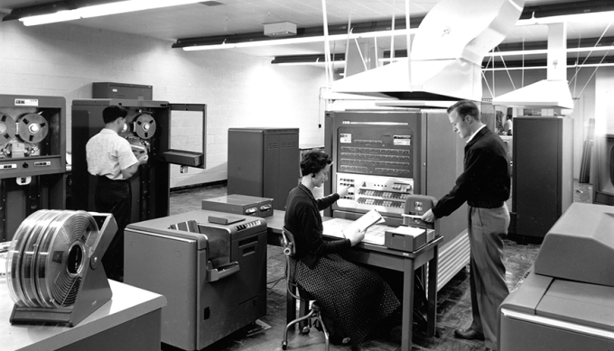 black-and-white photo of three people operating the computer in a machine room equipped with cooling ducts and tape reels