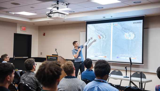 Stefan at a podium gesturing to his presentation on the screen in front of a seated audience